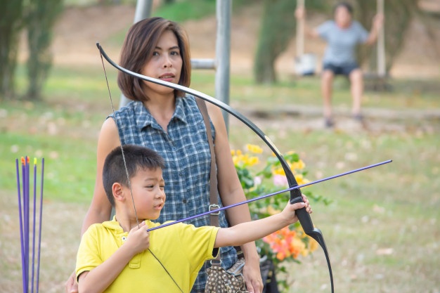 Asian boys holding a bow in camp adventure and the woman standing behind background blurry tree.